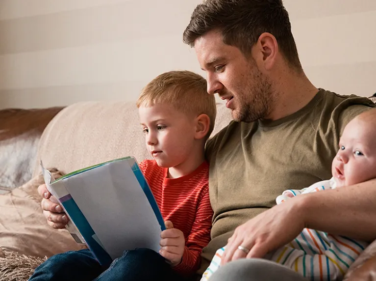 A photo of a father sitting with his son reading a book whilst holding a baby in the family's new build home.